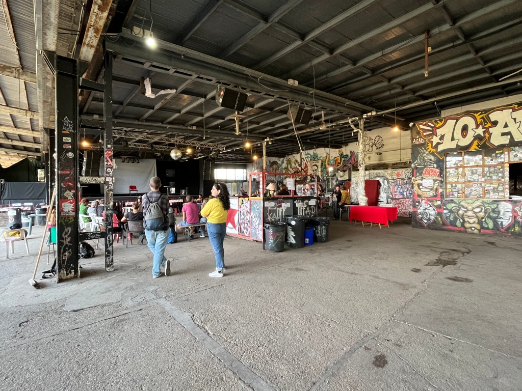 Photo depicting the inside canteen space of Acrobax. The walls are covered in graffiti art and the bar car be seen to the far right of the photo. In the centre stands a sound and lights area (I believe) and in the background to the right there is a central discussion taking place, with people gathered around, either seated or standing listening to the main speaker. Two lovely legends I met on the day stand in the mid ground to the right, chatting to each other. 